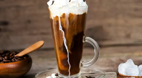 Ein erfrischender iced espresso mit Sahne, Marshmallows und Schokostreuseln, daneben Kaffeebohnen und Eiswürfel.