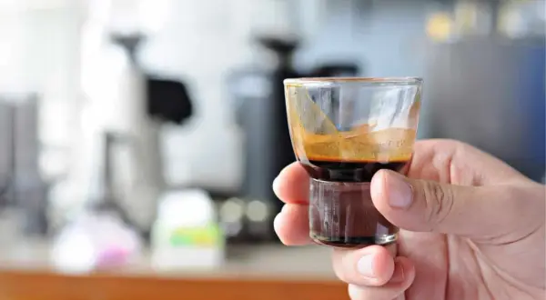 Eine Hand hält ein kleines Glas mit einem dunklen, cremigen Ristretto. Die Zubereitung ist perfekt, die Crema glänzt.