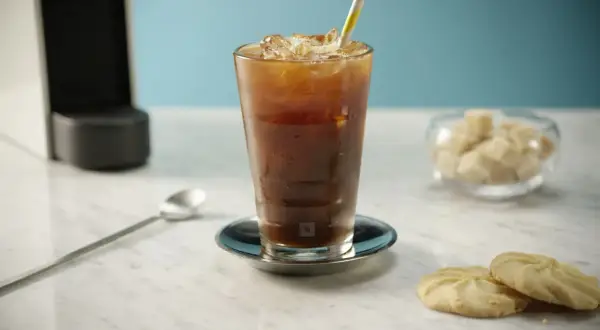 Erfrischender Nespresso Iced Coffee mit Eiswürfeln und einem bunten Strohhalm, daneben Kekse und Zuckerwürfel.