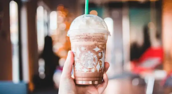 Eine Hand hält einen Schokoladen-Frappuccino von Starbucks, der mit Schlagsahne und einem grünen Strohhalm gekrönt ist. Perfekt für heiße Tage, diese Starbucks Rezepte Frappuccino sind ein Genuss.