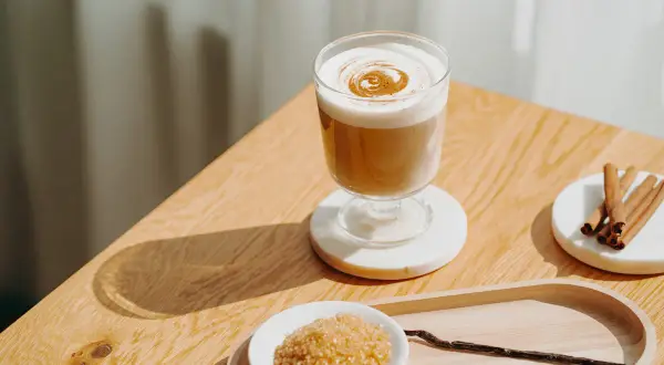 Ein cremiger Zimt Latte mit aufgeschäumter Milch und Zimtdekor. Daneben brauner Zucker, Zimtstangen und eine Vanilleschote.