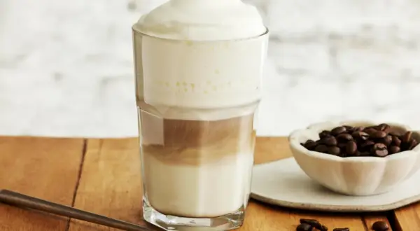 Ein hoher Latte Macchiato Eiskaffee mit Milchschaum, daneben eine Schale Kaffeebohnen und ein Löffel.