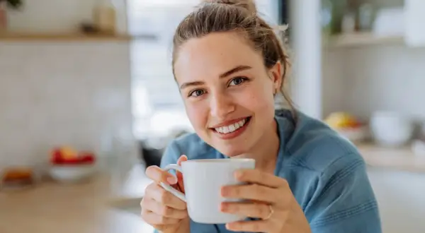 Lächelnde Frau hält eine Tasse Kaffee. Sie genießt den Moment, vielleicht trinkt sie alten Kaffee, um den Tag zu beginnen.