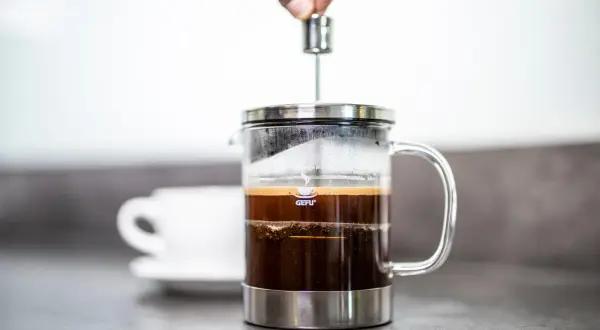 Eine Hand drückt den Stempel einer French Press herunter. So funktioniert eine French Press: Kaffee und heißes Wasser ziehen lassen, dann den Kaffeesatz abpressen.