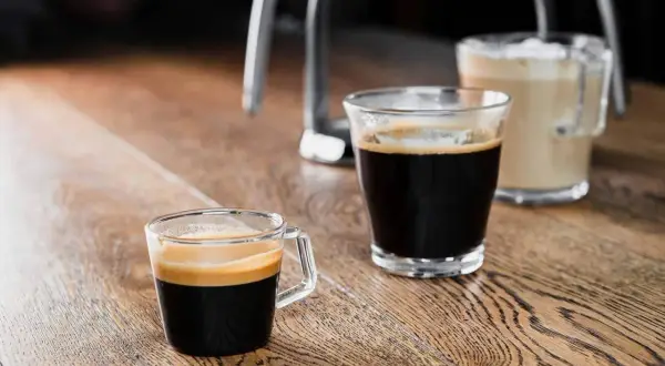 Zwei Gläser mit Kaffee stehen auf einem Holztisch. Ein kleiner starker Kaffee mit Crema im Vordergrund, daneben ein weiterer Kaffee und im Hintergrund ein Milchkaffee.