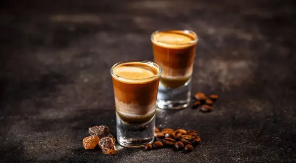 Zwei geschichtete Kaffeegetränke in Gläsern, umgeben von Kaffeebohnen und braunem Zucker. So kann man mit Kaffeebohnen köstliche Drinks zaubern.