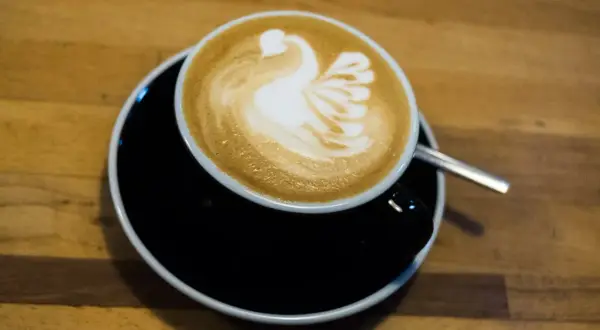 Kaffee mit einem Schwan aus Milchschaum zubereitet, serviert auf einem schwarzen Unterteller.