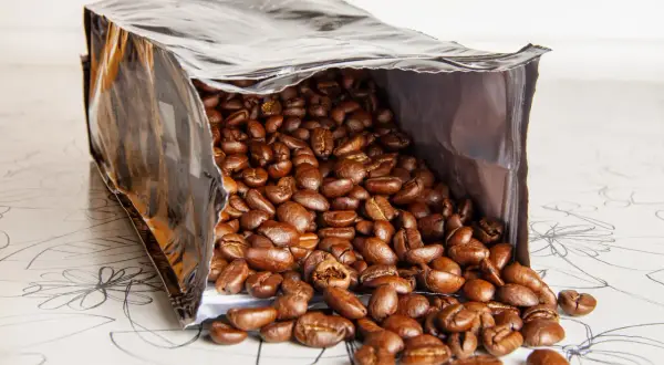 Ein offener Beutel mit gerösteten Kaffeebohnen, die auf eine gemusterte Oberfläche fallen. Die Frage, wie lange sind kaffeebohnen haltbar, beschäftigt jeden Kaffeeliebhaber.