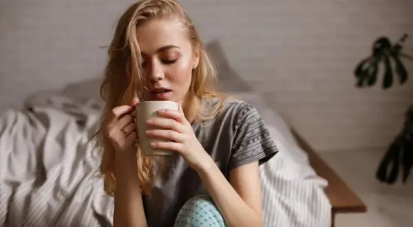 Junge Frau sitzt im Bett, hält eine Tasse und denkt nach. Ihr Kaffee schmeckt nicht mehr.