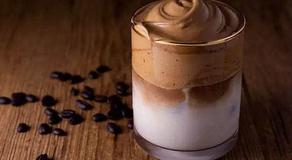 Ein cremiger Dalgona Kaffee ohne Zucker, serviert in einem Glas auf einem Holztisch, umgeben von Kaffeebohnen.