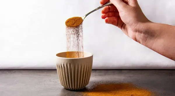 Eine Hand schüttet mit einem Löffel braunes Pulver in eine gerippte Tasse. Der Pulverregen erinnert an instant kaffee koffein.