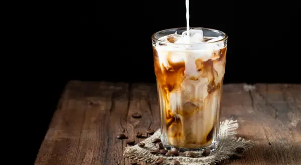 Milch wird in ein Glas mit Eis und Kaffee gegossen. Ein erfrischender Eiskaffee auf einem Holztisch.