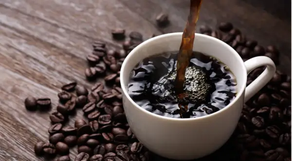 Heißer schwarzer Kaffee wird in eine weiße Tasse gegossen, umgeben von gerösteten Kaffeebohnen. Ein gesunder Start in den Tag.