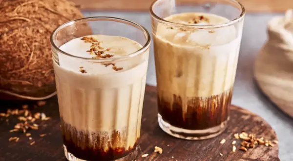 Zwei Gläser mit cremigem Kaffee mit Kokosmilch, garniert mit Kokosraspeln. Daneben eine Kokosnuss.