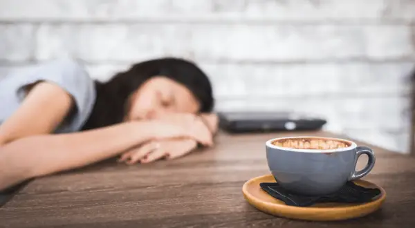 Eine Frau schläft auf dem Tisch, ihr Kopf ruht auf ihren Armen. Neben ihr steht eine Tasse Kaffee, die sie wohl abends trinken wollte.