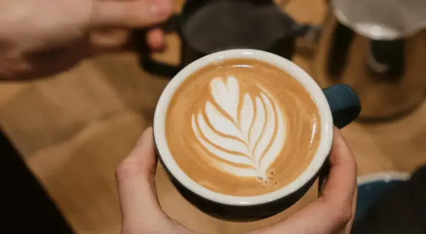 Perfekte Cappuccino Zubereitung: Eine Hand hält die Tasse mit Latte Art, während im Hintergrund Milch aufgeschäumt wird.