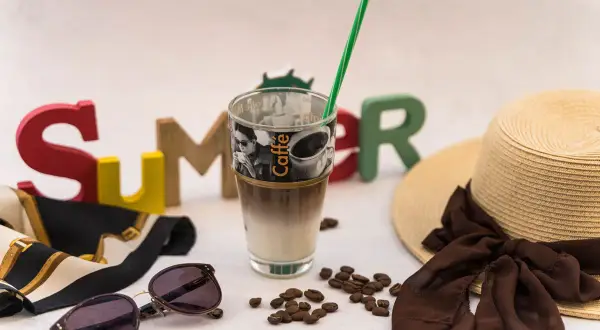 Ein erfrischender Vanilla Iced Coffee steht bereit für einen sonnigen Sommertag, umgeben von bunten Buchstaben, einer Sonnenbrille und einem Strohhut.
