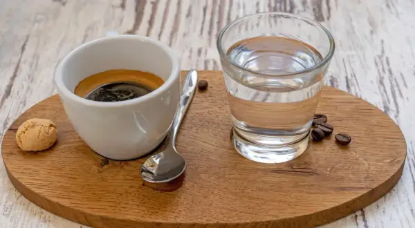 Ein Espresso mit einem Glas Wasser, einem Keks und Kaffeebohnen. Das Wasser ist wichtig, um den Gaumen zu reinigen und den vollen Geschmack des Espressos zu genießen.