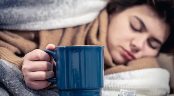 Müde Frau mit Fieber hält eine Tasse Kaffee, um sich aufzuwärmen. Medikamente liegen daneben.