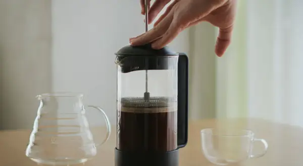 Hände drücken den Stempel einer French Press herunter, um Kaffee zu kochen. Daneben stehen eine leere Glaskaraffe und eine Tasse.
