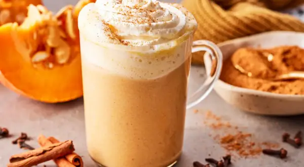 Ein cremiger Pumpkin Spice Latte mit Sahne und Zimt, daneben Kürbisspalten, Zimtstangen und Gewürznelken.