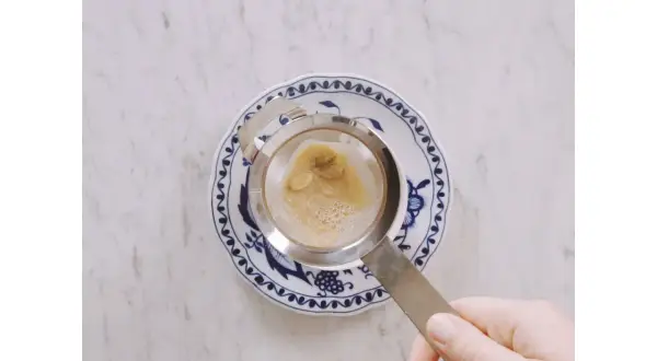 Hand hält Sieb mit braunem Schaum über blau-weißem Teller. Ein leckerer soy latte wird zubereitet.