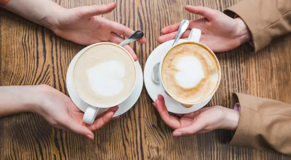 Zwei Hände reichen sich zwei Kaffees, ein Flat White und ein Cappuccino, mit Herz-Latte-Art auf Holztisch.