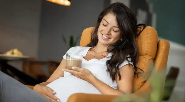 Glückliche Schwangere genießt einen Kaffee. Sie sitzt entspannt in einem Sessel und hält ein Glas mit einem cremigen Getränk.