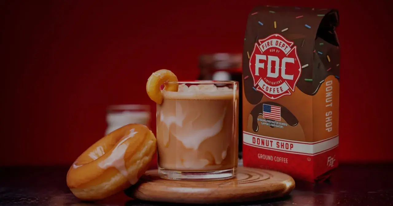 Ein cremiger iced americano mit einem Donut-Garnitur, daneben ein Donut und eine Packung Fire Dept. Coffee.
