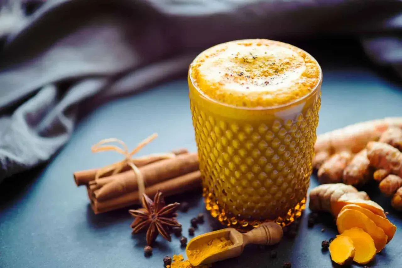 Ein goldgelbes Getränk mit Kurkuma in Kaffee, verfeinert mit Zimt und Sternanis. Daneben liegen frische Kurkumawurzeln und eine kleine Holzschaufel mit Kurkumapulver.