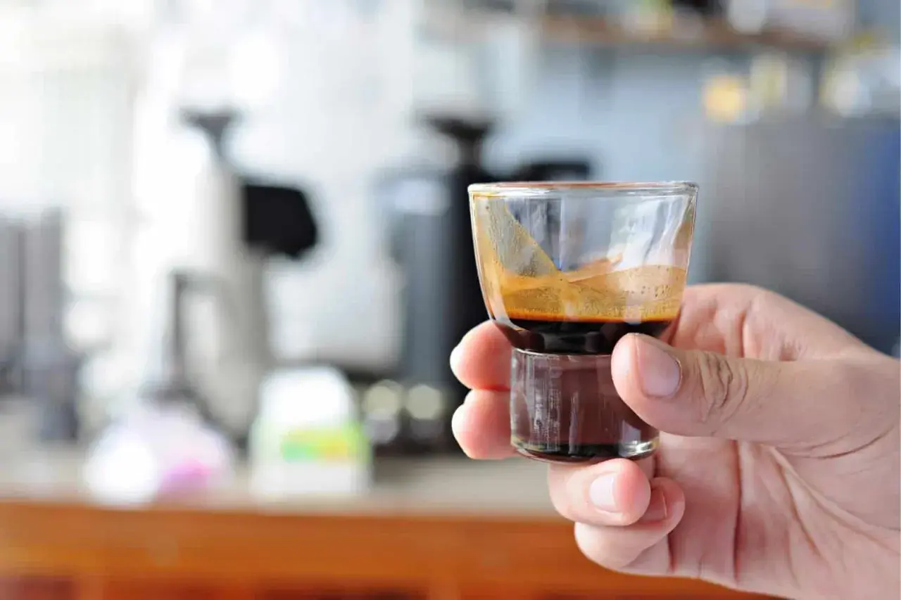 Eine Hand hält ein kleines Glas mit einem dunklen, cremigen Ristretto. Die Zubereitung ist perfekt, die Crema glänzt.