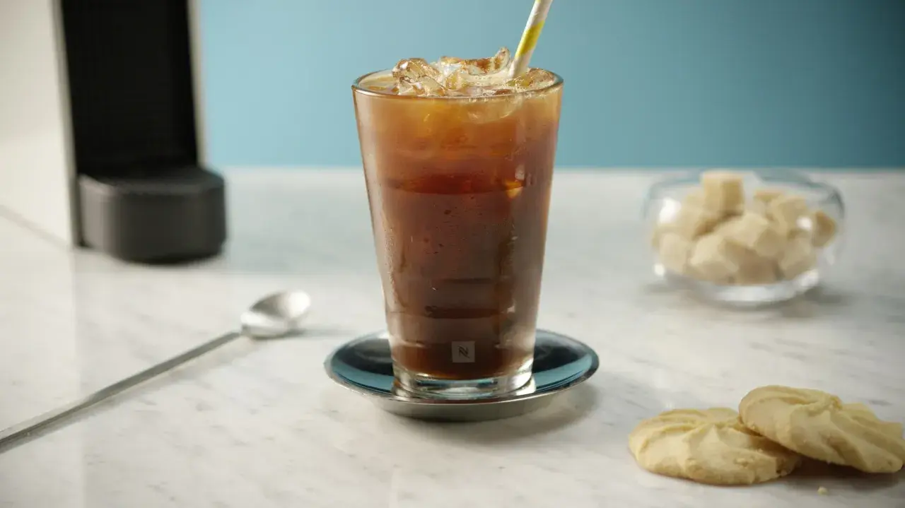 Erfrischender Nespresso Iced Coffee mit Eiswürfeln und einem bunten Strohhalm, daneben Kekse und Zuckerwürfel.