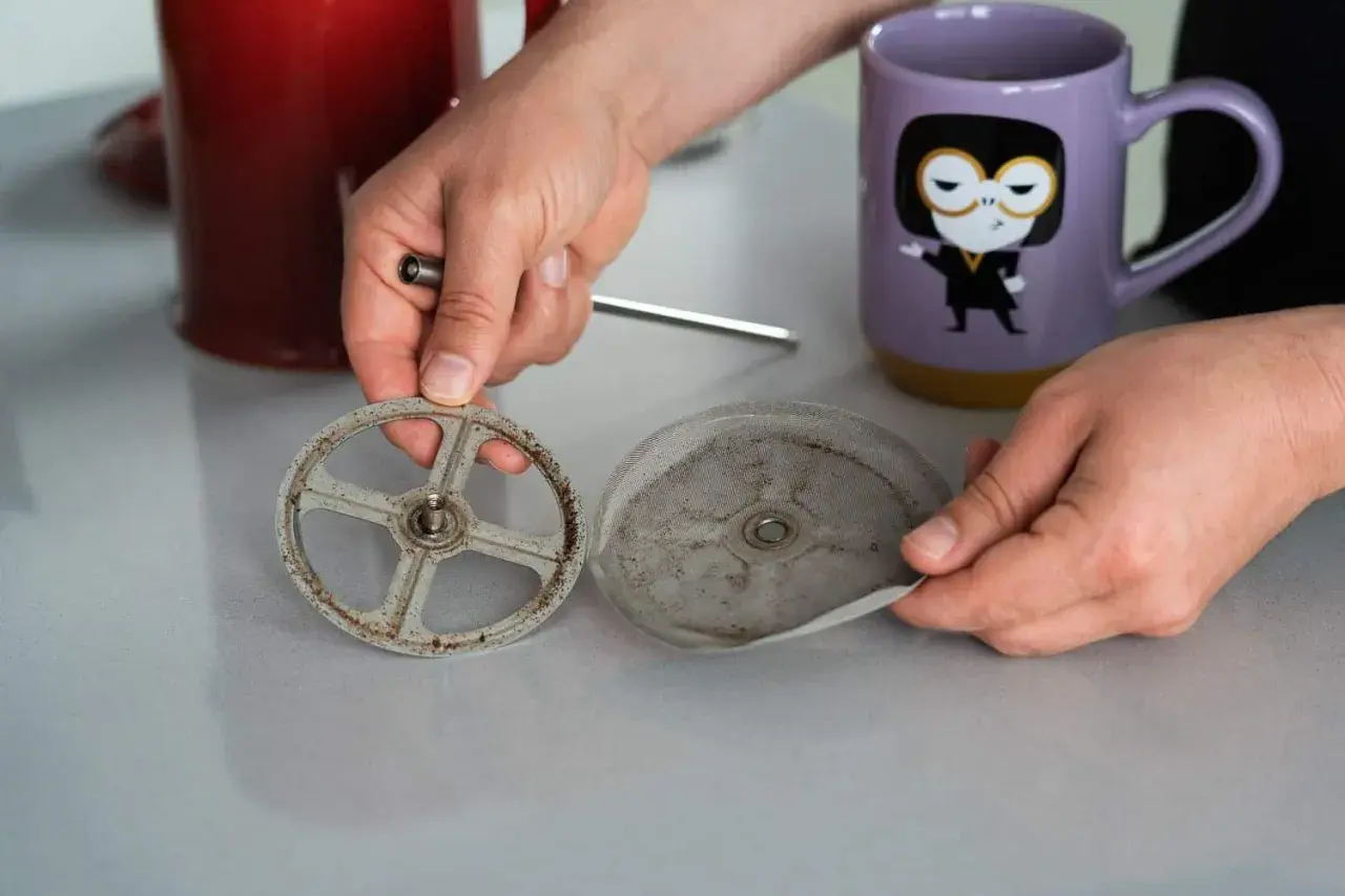 Hände zerlegen eine schmutzige French Press, um sie zu reinigen. Der Siebträger und das Sieb sind mit Kaffeesatz bedeckt.