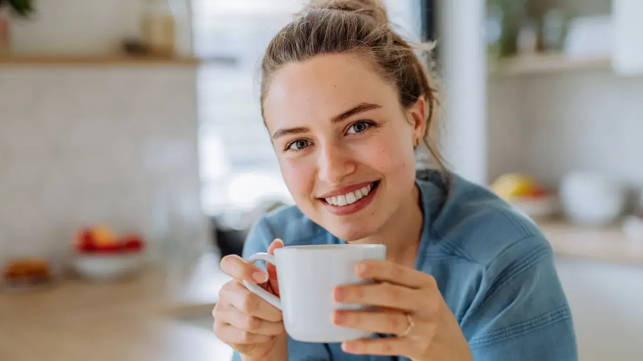 Lächelnde Frau hält eine Tasse Kaffee. Sie genießt den Moment, vielleicht trinkt sie alten Kaffee, um den Tag zu beginnen.