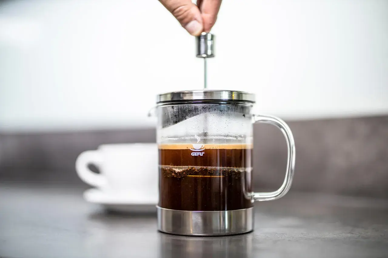 Eine Hand drückt den Stempel einer French Press herunter. So funktioniert eine French Press: Kaffee und heißes Wasser ziehen lassen, dann den Kaffeesatz abpressen.