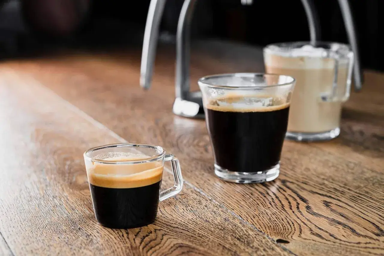 Zwei Gläser mit Kaffee stehen auf einem Holztisch. Ein kleiner starker Kaffee mit Crema im Vordergrund, daneben ein weiterer Kaffee und im Hintergrund ein Milchkaffee.