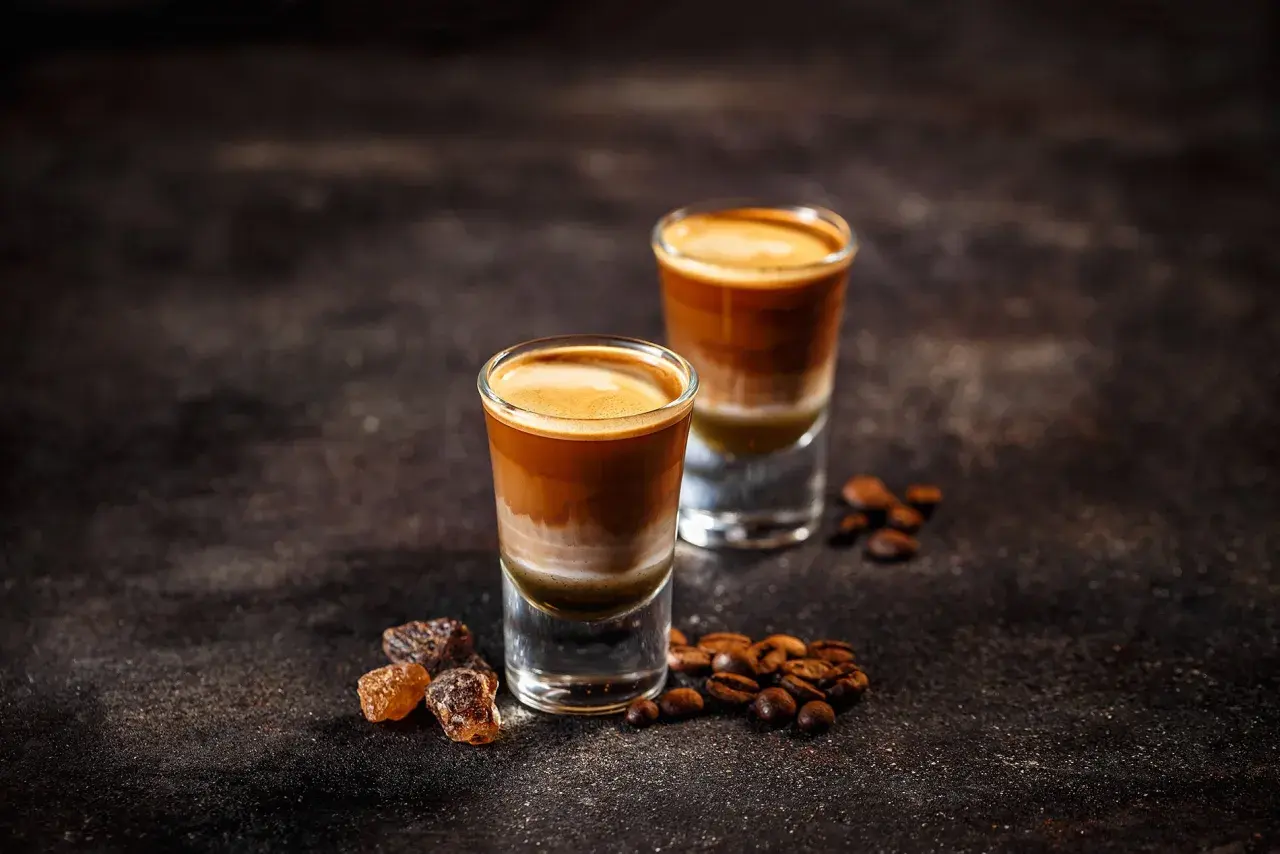 Zwei geschichtete Kaffeegetränke in Gläsern, umgeben von Kaffeebohnen und braunem Zucker. So kann man mit Kaffeebohnen köstliche Drinks zaubern.