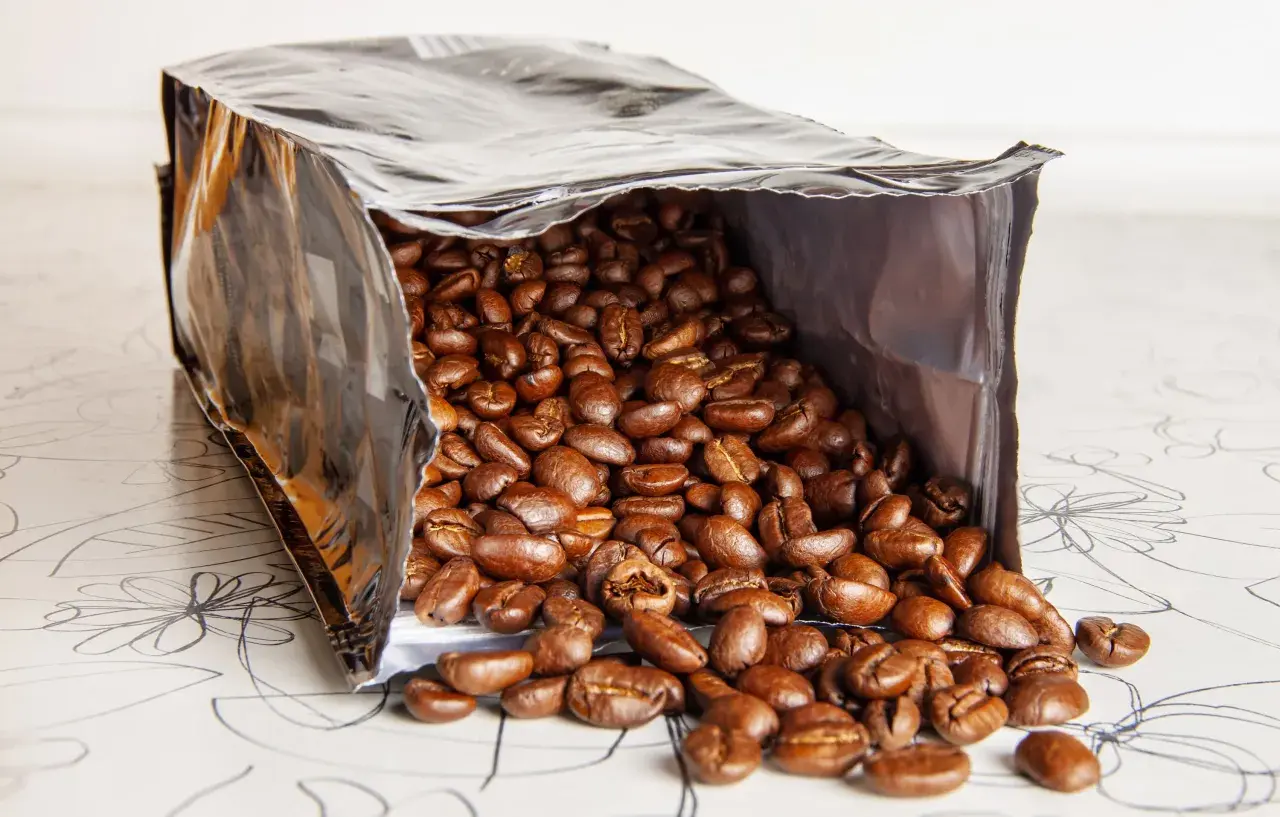 Ein offener Beutel mit gerösteten Kaffeebohnen, die auf eine gemusterte Oberfläche fallen. Die Frage, wie lange sind kaffeebohnen haltbar, beschäftigt jeden Kaffeeliebhaber.