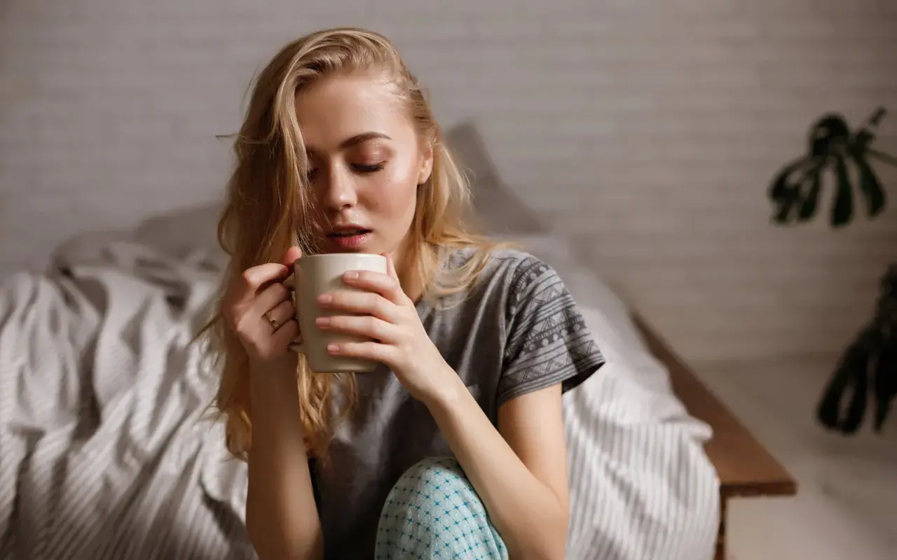 Junge Frau sitzt im Bett, hält eine Tasse und denkt nach. Ihr Kaffee schmeckt nicht mehr.
