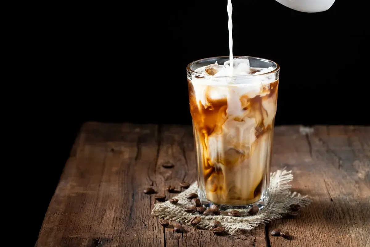 Milch wird in ein Glas mit Eis und Kaffee gegossen. Ein erfrischender Eiskaffee auf einem Holztisch.