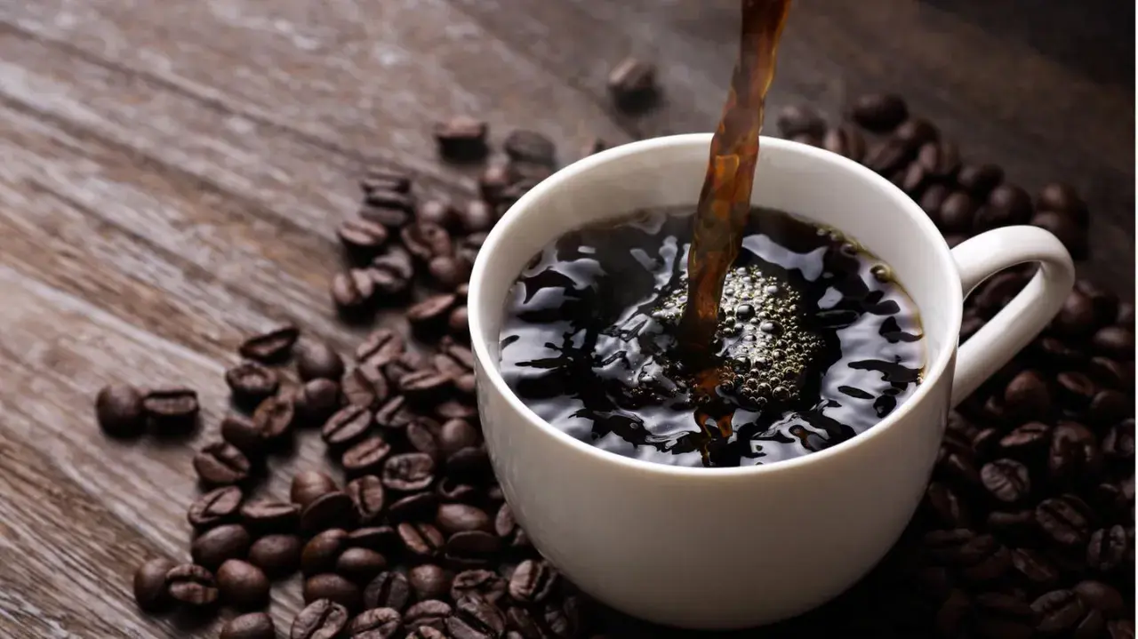 Heißer schwarzer Kaffee wird in eine weiße Tasse gegossen, umgeben von gerösteten Kaffeebohnen. Ein gesunder Start in den Tag.