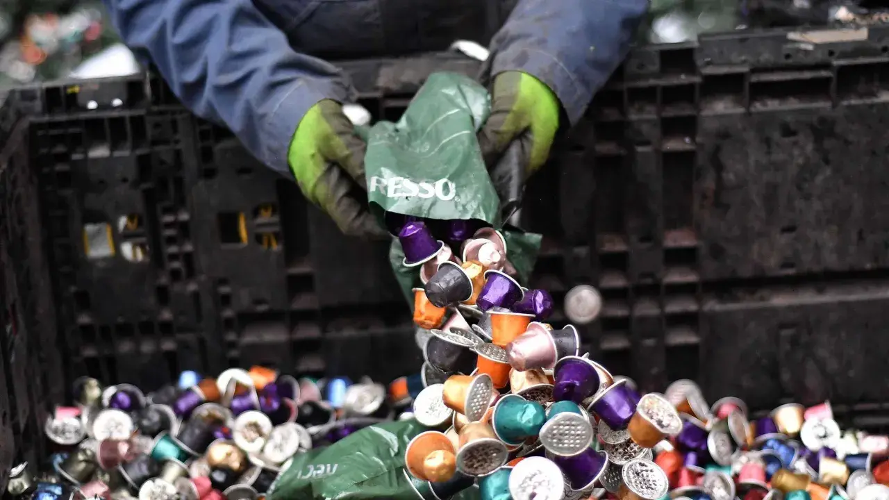 Hände in Handschuhen kippen bunte Nespresso-Kapseln aus einer grünen Tüte in einen schwarzen Behälter. Die fachgerechte Entsorgung von Nespresso-Kapseln ist wichtig.
