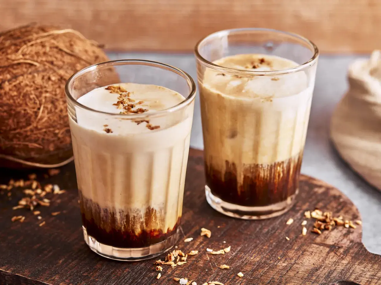 Zwei Gläser mit cremigem Kaffee mit Kokosmilch, garniert mit Kokosraspeln. Daneben eine Kokosnuss.