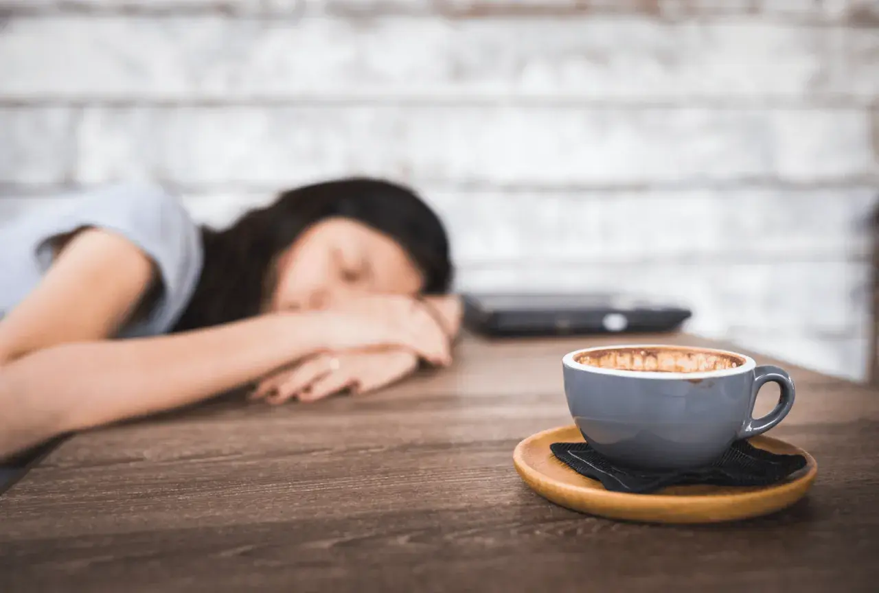 Eine Frau schläft auf dem Tisch, ihr Kopf ruht auf ihren Armen. Neben ihr steht eine Tasse Kaffee, die sie wohl abends trinken wollte.