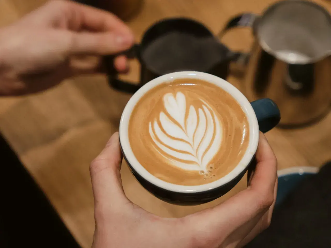 Perfekte Cappuccino Zubereitung: Eine Hand hält die Tasse mit Latte Art, während im Hintergrund Milch aufgeschäumt wird.