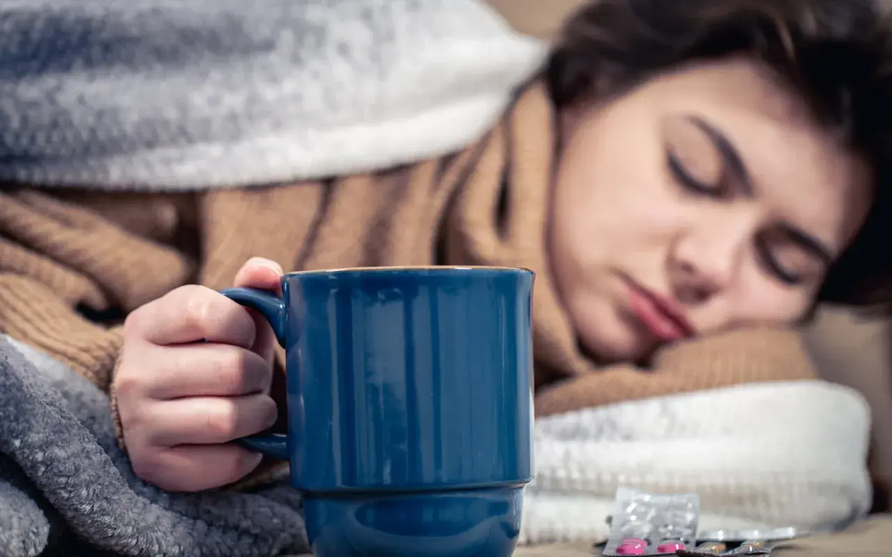 Müde Frau mit Fieber hält eine Tasse Kaffee, um sich aufzuwärmen. Medikamente liegen daneben.