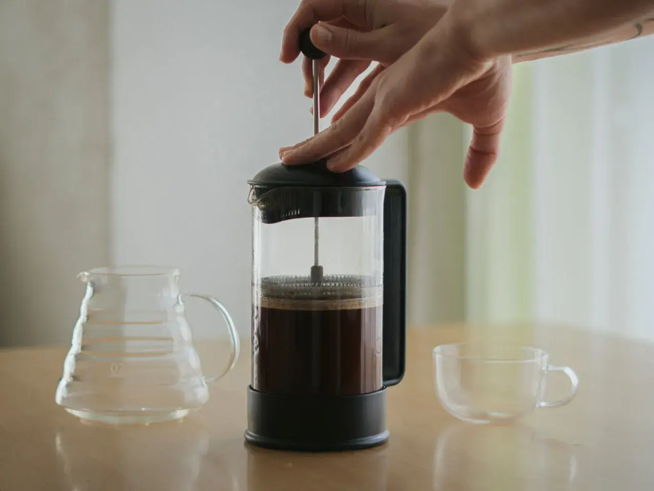 Hände drücken den Stempel einer French Press herunter, um Kaffee zu kochen. Daneben stehen eine leere Glaskaraffe und eine Tasse.
