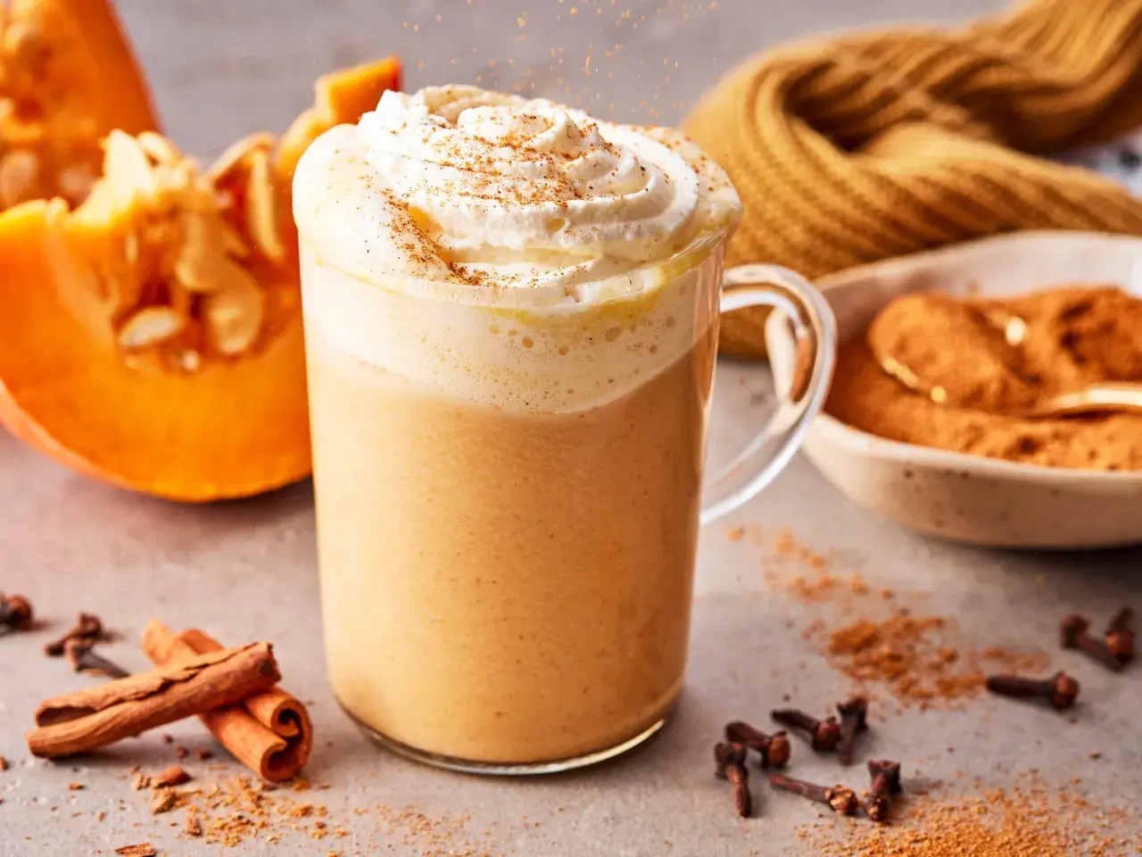 Ein cremiger Pumpkin Spice Latte mit Sahne und Zimt, daneben Kürbisspalten, Zimtstangen und Gewürznelken.