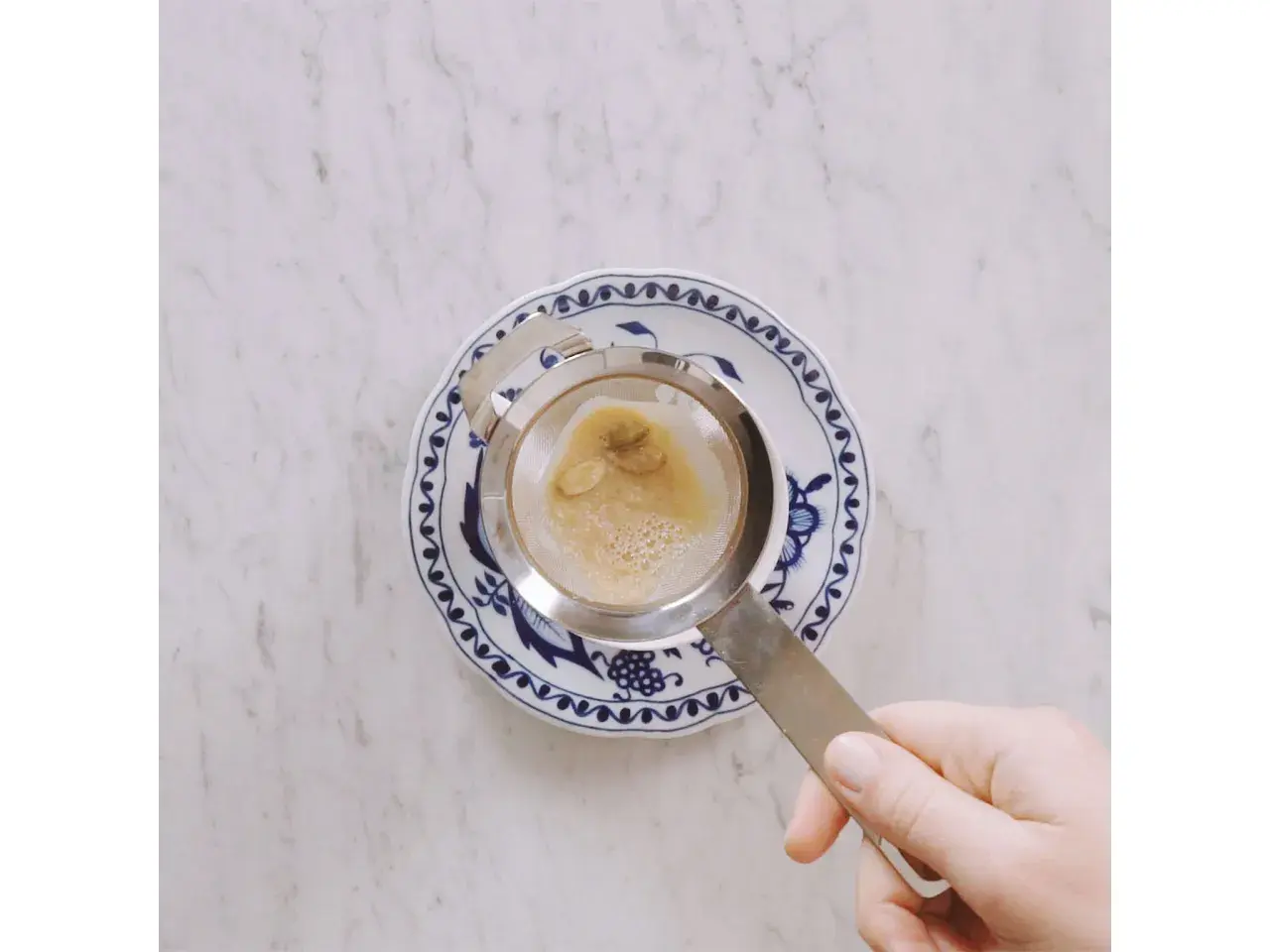 Hand hält Sieb mit braunem Schaum über blau-weißem Teller. Ein leckerer soy latte wird zubereitet.