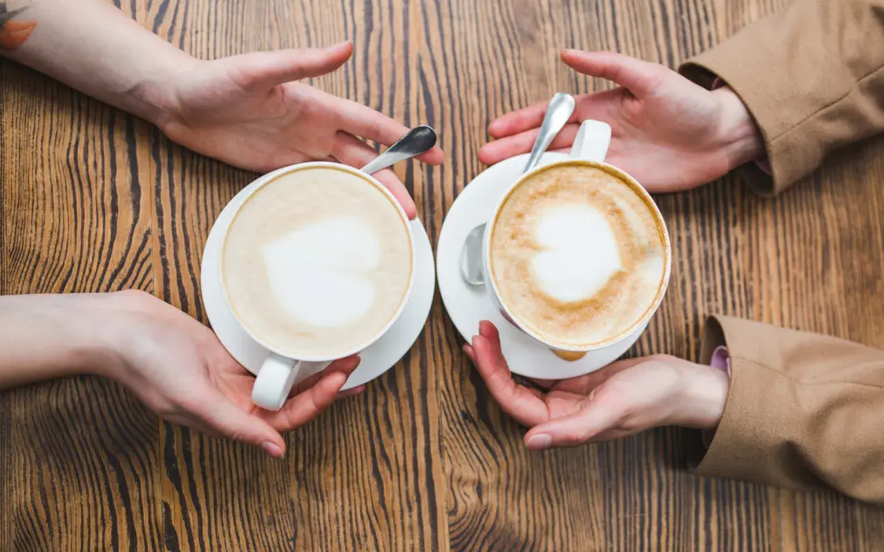 Zwei Hände reichen sich zwei Kaffees, ein Flat White und ein Cappuccino, mit Herz-Latte-Art auf Holztisch.